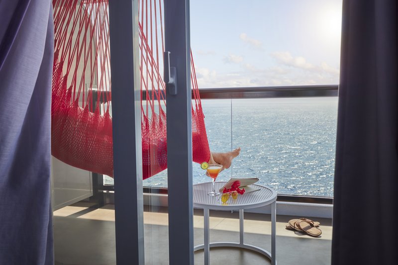 View from inside a Virgin Voyages Sea Terrace cabin through the open balcony doors: red rope hammock with someone's feet up, sandals, a cocktail on a round side table, and open ocean beyond