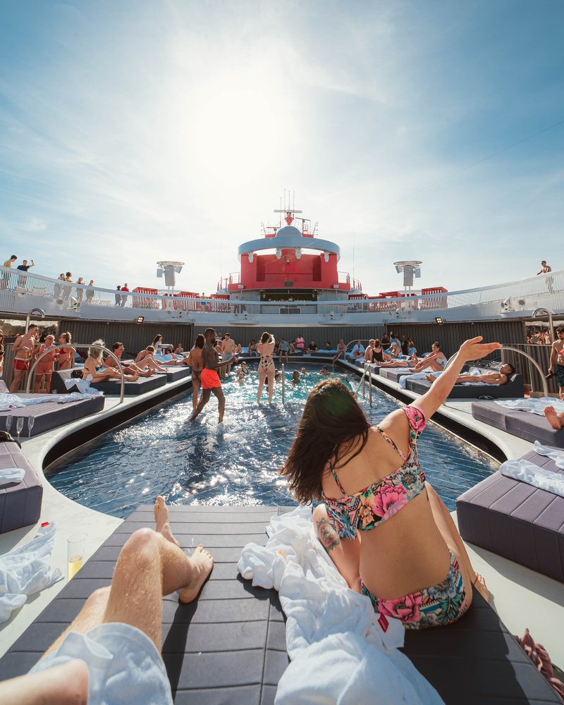Adult Sailors sitting at the edge of the Aquatic Club pool on a Virgin Voyages ship, sun overhead, red smokestack and curved sun loungers in the background