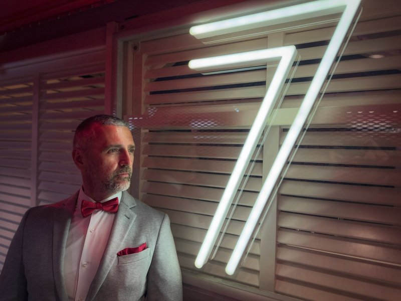 Igor Fedulov on a Virgin Voyages ship, grey blazer with red bow tie and pocket square, lit by red ambient light and a neon 77 backdrop