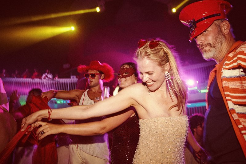 Scarlet Night on a Virgin Voyages ship: dancers in red captain hats, masquerade masks, and feather headpieces moving on the pool-deck dance floor under purple and yellow stage lighting