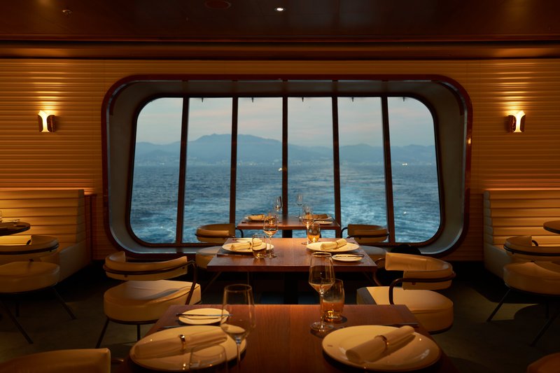 The Wake dining room aboard a Virgin Voyages ship: wicker chairs and dressed tables facing a stern window framing ocean and distant mountains at dusk